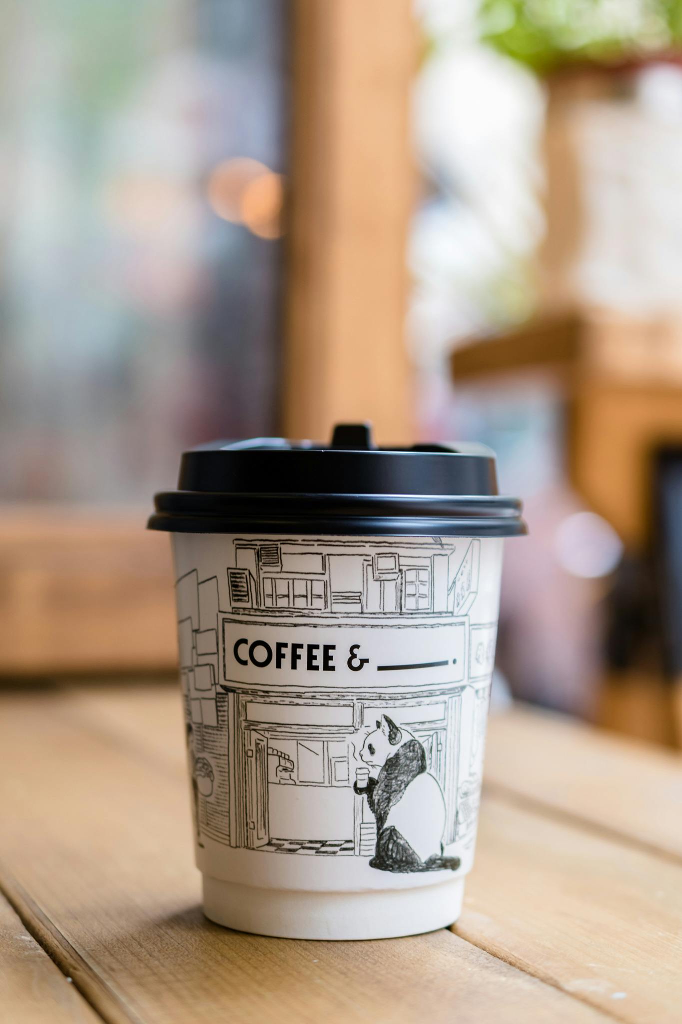 Paper coffee cup with black lid on a wooden surface, featuring a panda illustration and text.