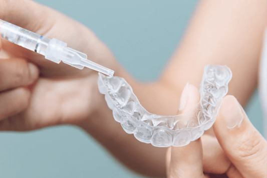 A person applying gel from a syringe to a clear dental aligner held in their hand, set against a soft background.